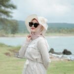 A woman in a hijab and sunglasses poses elegantly by the coast in Indonesia.